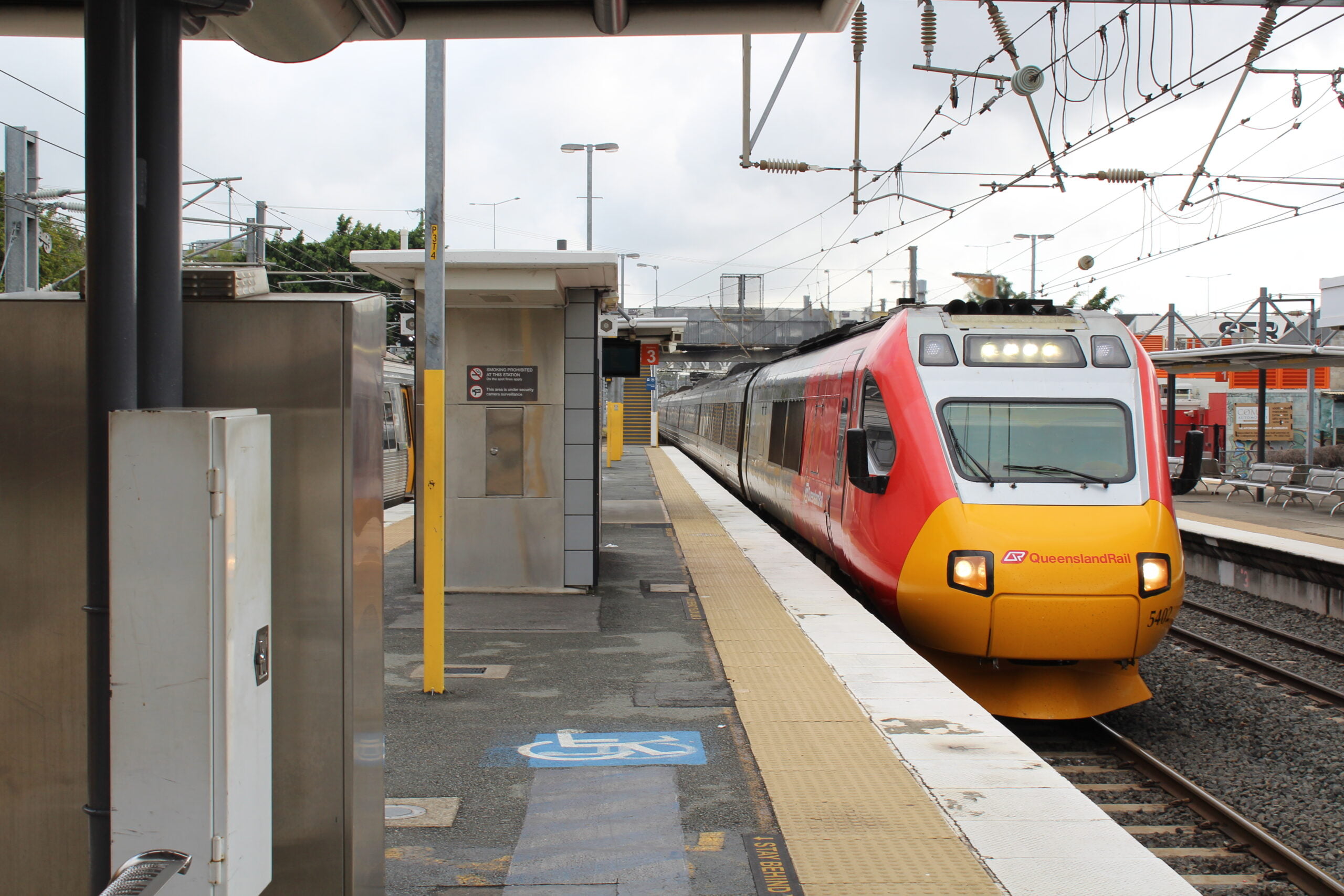 A stationary train with a Queensland Rail logo printed at the front, is shown on the tracks, positioned at the platform of a station.